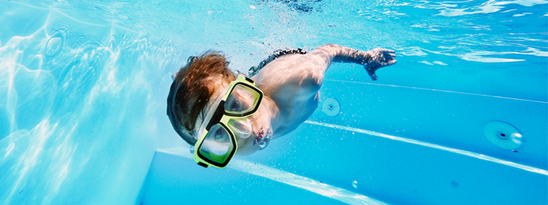 Under vattnet i en pool med medelhavsblått vatten simmar en pojke med dykarglasögon. Ur munnen bubblar det.